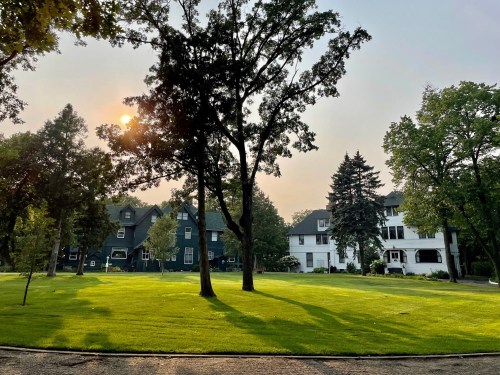 A serene park with green grass, tall trees, and two houses in the background, illuminated by a soft sunset glow.