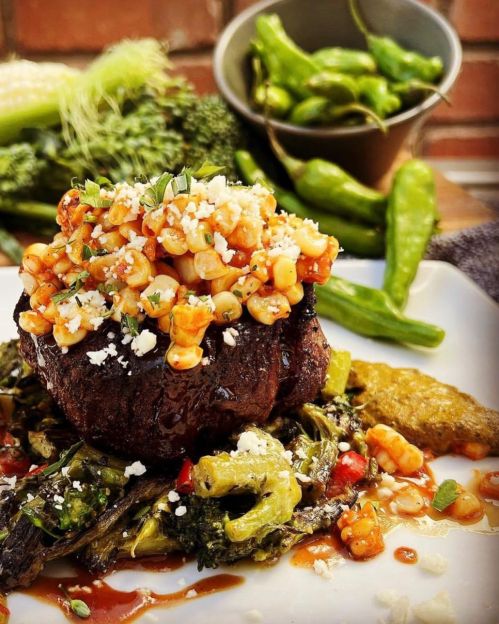 A gourmet dish featuring a grilled steak topped with corn salsa, served with vegetables and a side of green peppers.