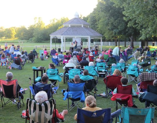 A large crowd sits in folding chairs facing a gazebo during an outdoor event, surrounded by trees and a grassy field.