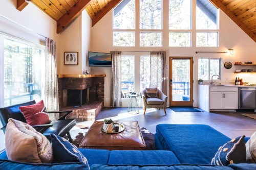Cozy living room with high ceilings, large windows, a fireplace, and modern furnishings in a forested setting.