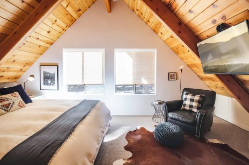 Cozy attic bedroom with wooden beams, large windows, a black chair, and a TV, featuring a warm, inviting atmosphere.