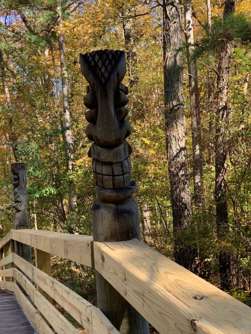 Wooden totem poles stand on a wooden walkway surrounded by colorful autumn trees.