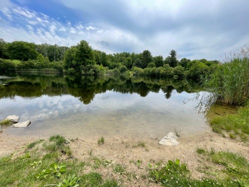 A serene lake surrounded by lush greenery and trees, reflecting the cloudy sky above.