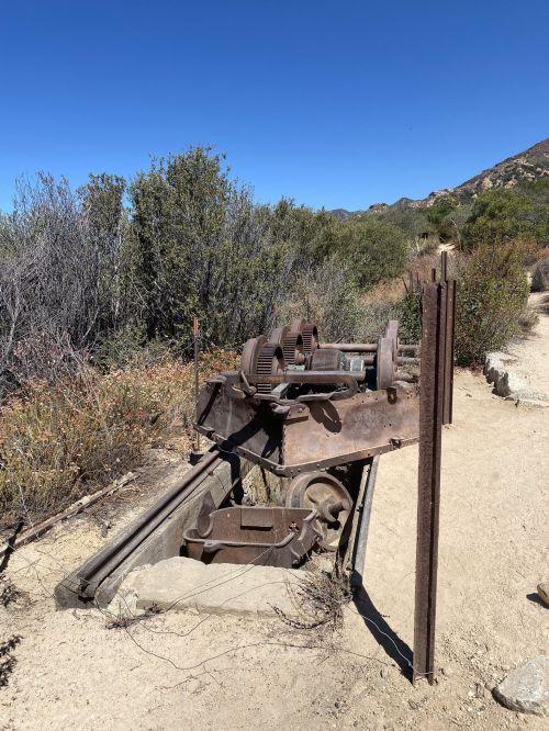 An old, rusted mining machine sits on a dirt path surrounded by shrubs and mountains under a clear blue sky.
