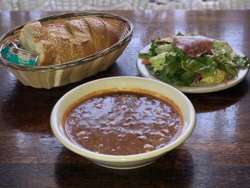 A bowl of chili, a side salad, and a basket of sliced bread on a wooden table.