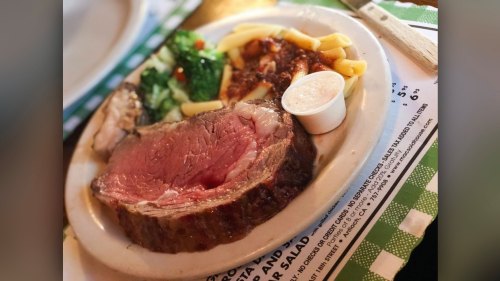 A plate featuring a slice of prime rib, pasta, vegetables, and a small cup of sauce.