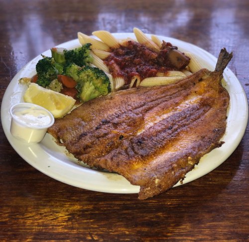 A plate featuring a fried fish fillet, pasta with sauce, steamed broccoli, a lemon wedge, and a side of dipping sauce.