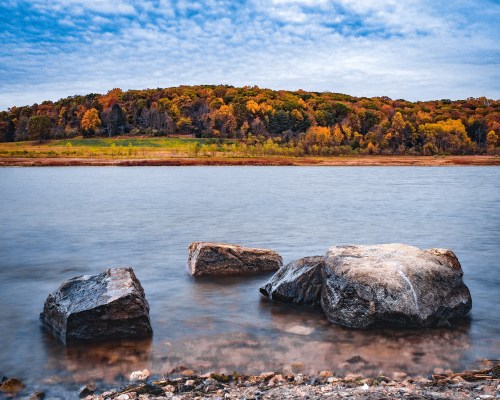 A serene river scene with smooth rocks in the foreground and a colorful, tree-covered hill in the background.