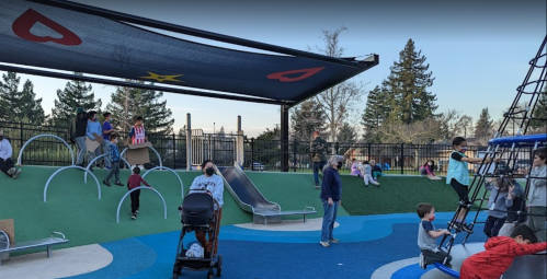 Children play on a colorful playground with slides and climbing structures, while adults supervise nearby.