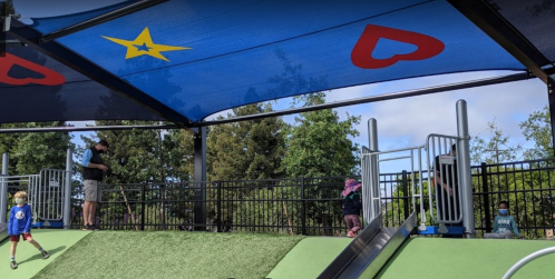 Children playing on a playground under a colorful canopy with stars and hearts, surrounded by trees.