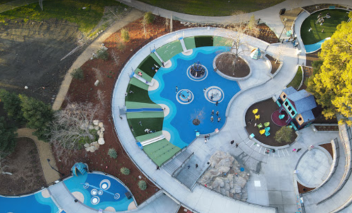Aerial view of a colorful water play area with slides, fountains, and grassy spaces, surrounded by trees.