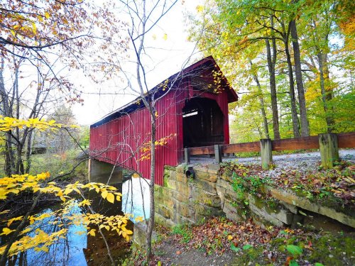 A red covered bridge spans a small stream, surrounded by autumn foliage and trees.