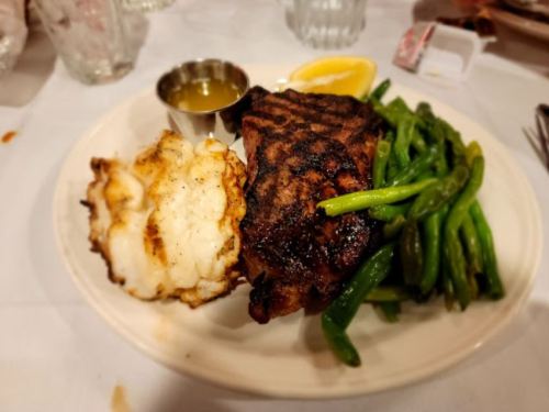 A plate with grilled steak, green beans, and a side of lobster tail, garnished with lemon and dipping sauce.