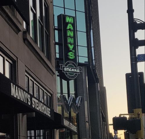 Sign for Manny's Steakhouse illuminated in green, located on a modern building with a city street view.