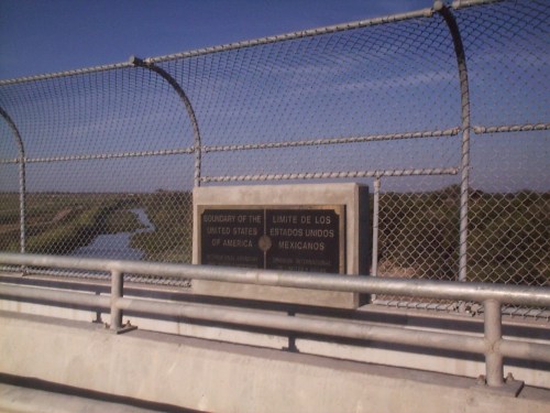 A fence along a bridge with a sign marking the border between the United States and Mexico, overlooking a river.