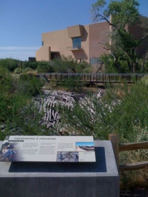 A sign about preservation in front of a dry creek bed, with a modern building and trees in the background.