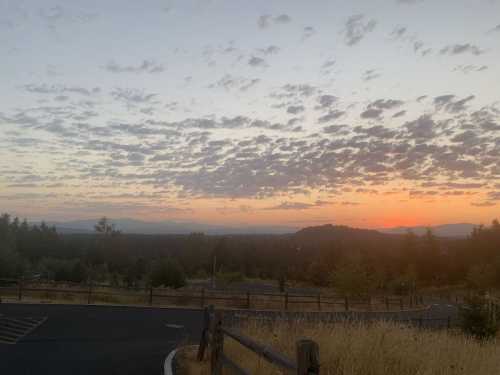 A serene sunset over a landscape with rolling hills, scattered clouds, and a wooden fence in the foreground.