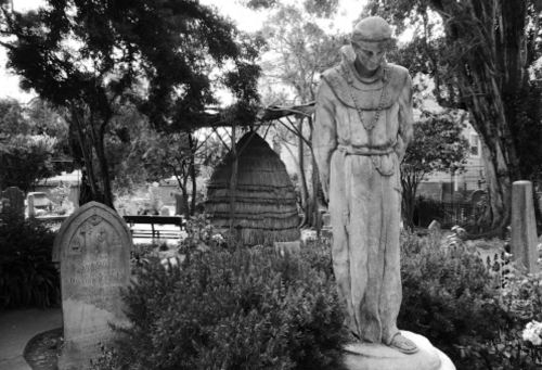 A somber statue stands in a cemetery, surrounded by greenery and gravestones, evoking a sense of reflection.