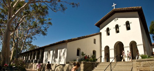 Historic building with a cross, featuring arched doorways and a bell tower, surrounded by trees and people.