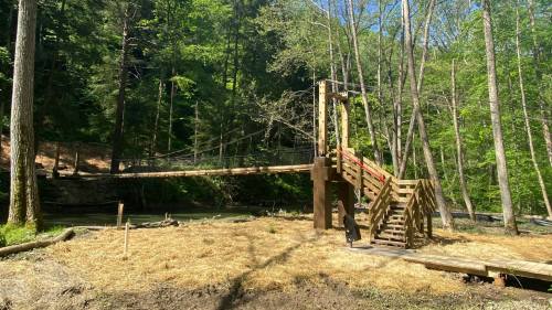 A wooden suspension bridge surrounded by trees, leading over a river with a grassy area in the foreground.