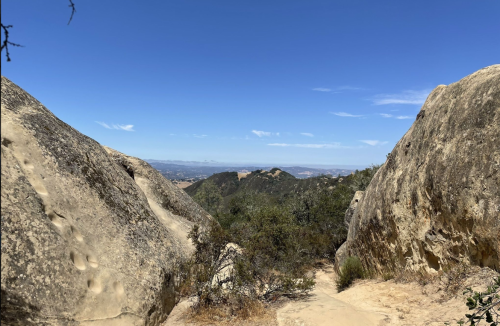 A scenic view between rocky formations, with a clear blue sky and distant mountains in the background.