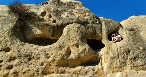 A couple sits in a rock formation resembling a face, with blue sky above and textured stone around them.
