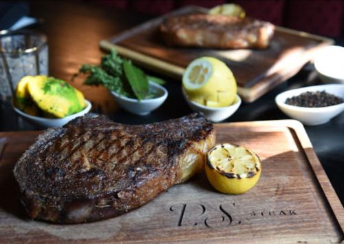 A beautifully grilled steak on a wooden board, accompanied by lemon halves and fresh herbs, with another steak in the background.