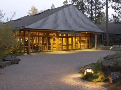 A modern building with large glass windows and wooden accents, surrounded by trees and a paved pathway.