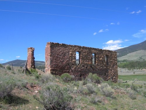Ruins of a stone building with windows, surrounded by grass and shrubs, against a backdrop of mountains and blue sky.