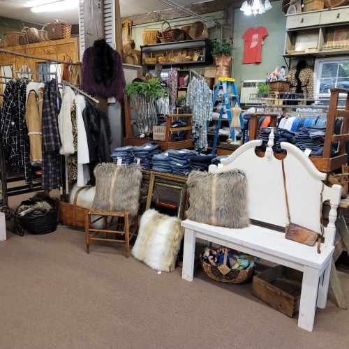 A cozy shop interior featuring clothing racks, denim stacks, and rustic furniture with decorative pillows and baskets.