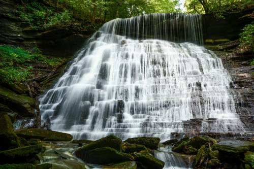 A serene waterfall cascading over rocky steps, surrounded by lush green foliage and moss-covered stones.