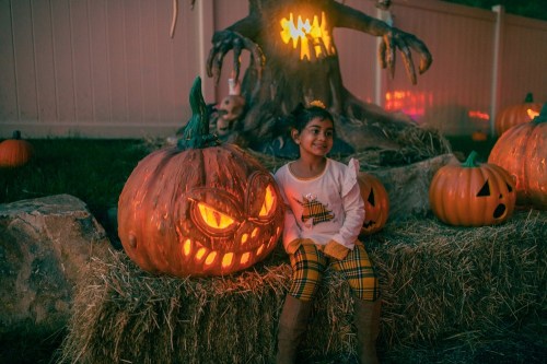 A child sits on hay next to a carved pumpkin with a mischievous face, surrounded by other pumpkins in a Halloween setting.
