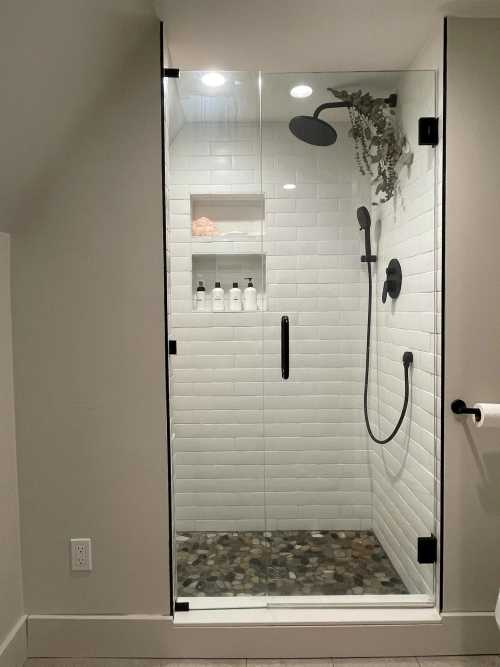 Modern glass shower with white tiles, a rainfall showerhead, and a pebble floor, featuring shelves for toiletries.