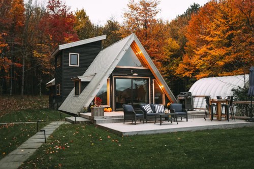A modern A-frame cabin surrounded by vibrant autumn foliage, featuring a patio with seating and a grill.
