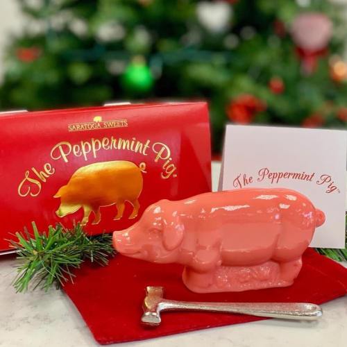 A pink peppermint pig candy sits on a red cloth, with a festive box and card in the background, against a Christmas tree.
