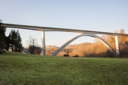 A modern bridge with a curved design spans over a grassy area, surrounded by trees and hills in the background.