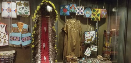Display of traditional Native American garments and art, including a feathered headdress and intricately designed bags.