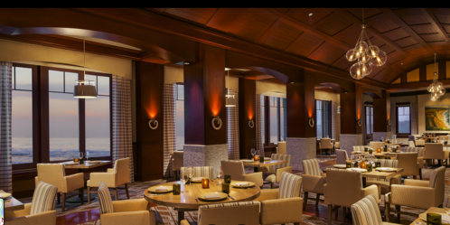 Elegant restaurant interior with wooden accents, soft lighting, and ocean views, featuring round tables and cozy seating.