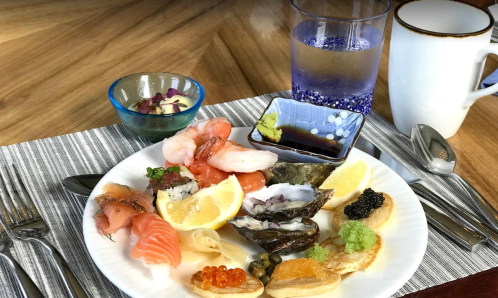 A beautifully arranged plate of sushi, seafood, and garnishes, accompanied by drinks and condiments on a table.