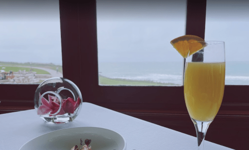 A glass of orange juice sits on a table with a flower arrangement, overlooking a scenic ocean view through a window.