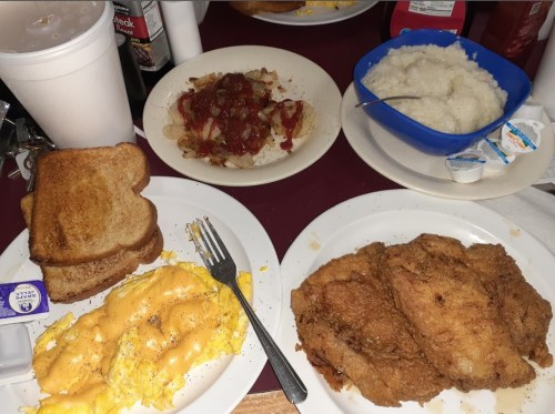 A table set with plates of fried fish, cheesy scrambled eggs, toast, grits, and a side of hash browns with ketchup.