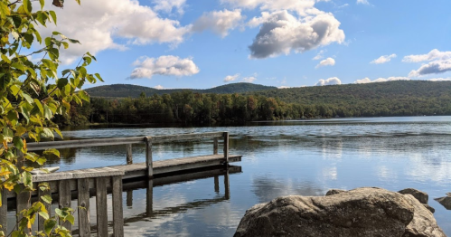 A serene lake view with a wooden dock, surrounded by trees and mountains under a blue sky with fluffy clouds.