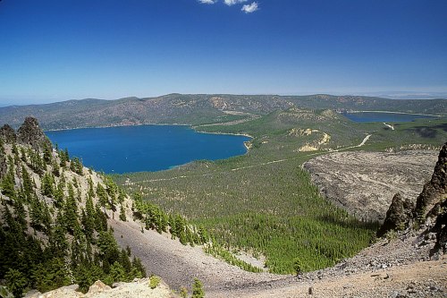A panoramic view of a blue lake surrounded by lush green forests and mountains under a clear blue sky.