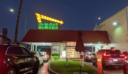 A brightly lit In-N-Out Burger drive-thru at night, with cars lined up and a moon visible in the sky.