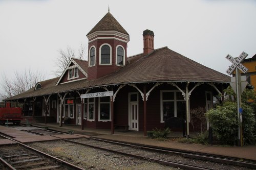 Historic train station with a distinctive tower, surrounded by tracks and greenery, under a cloudy sky.