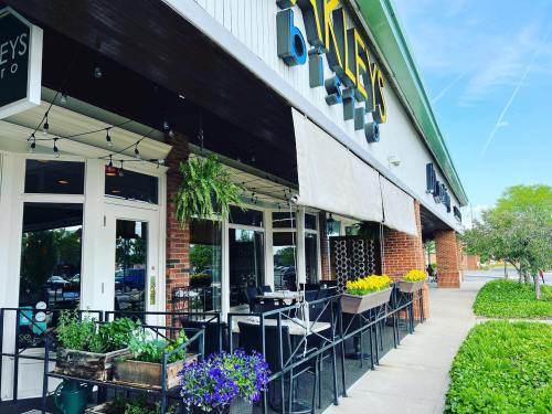 Exterior of a restaurant with outdoor seating, planters, and a sunny sky in the background.