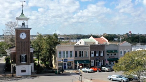 A scenic view of a clock tower and shops along a waterfront, with trees and a cloudy sky in the background.
