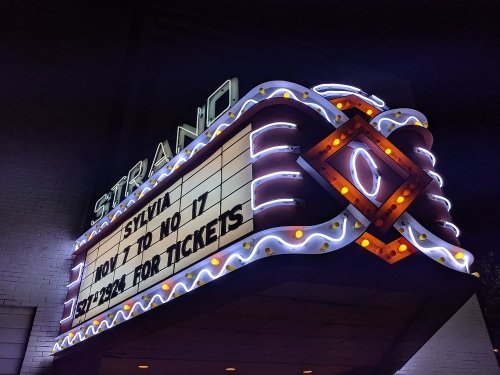 Neon-lit marquee of the Strano theater displaying "Sylvia" and "How to No It" with ticket information.