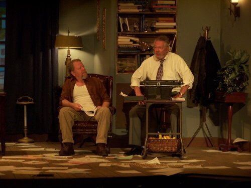Two men sit in a cluttered room, one in a chair and the other at a desk, engaged in a serious conversation.