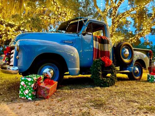 A vintage blue truck decorated for the holidays, surrounded by Christmas gifts and festive lights.